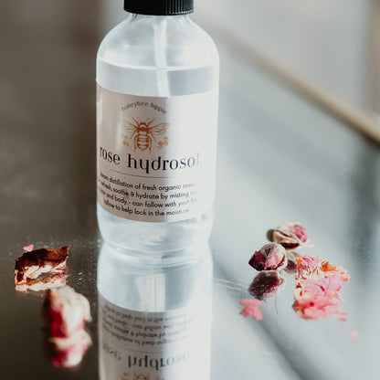 an image of a spray bottle of rose hydrosol spray sitting on a mirror next to some organic rose petals