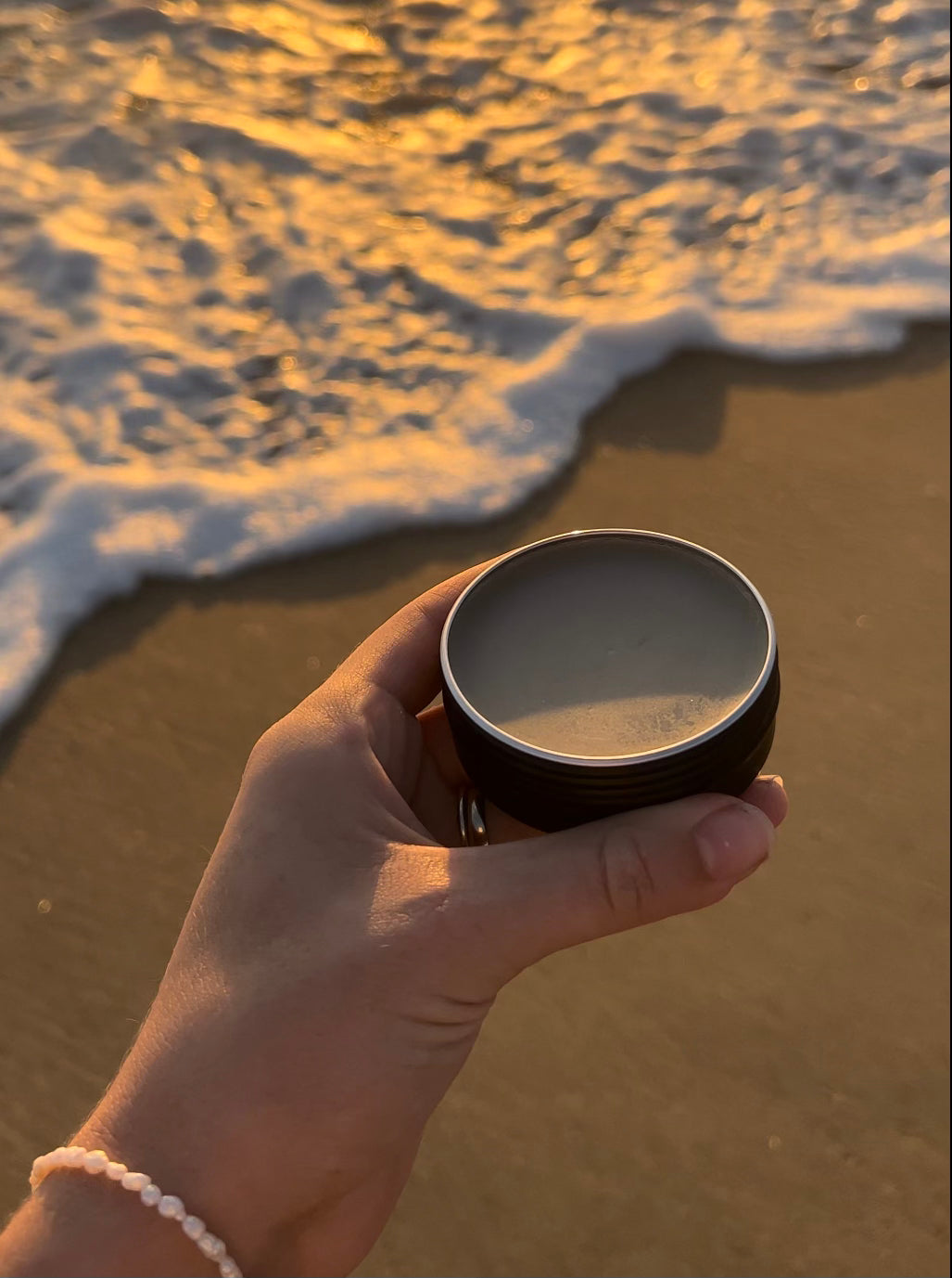a photo showing someone holding an open tin of miracle mud organic spot treatment natural skincare with sand & ocean in the background